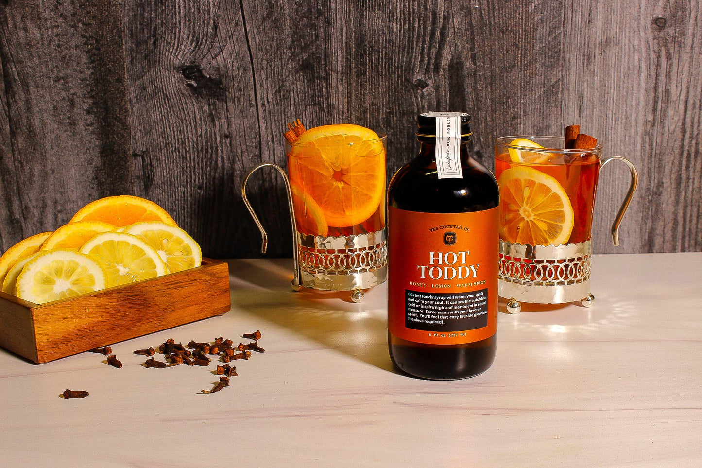 Bottle of Hot Toddy Syrup with glasses of hot toddy and citrus slices on a wooden surface.