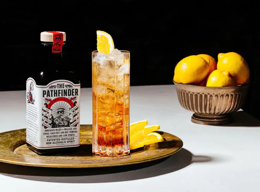 A bottle labeled 'The Pathfinder' with hemp and root depicted, alongside a glass filled with a amber-colored liquid and ice, garnished with an orange slice. Lemons are visible in the background.