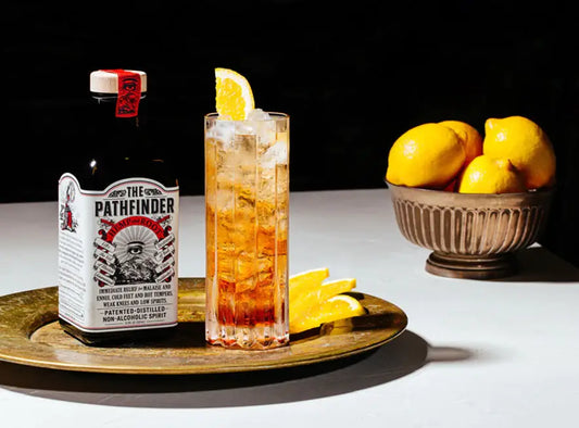 A bottle labeled 'The Pathfinder' with hemp and root depicted, alongside a glass filled with a amber-colored liquid and ice, garnished with an orange slice. Lemons are visible in the background.