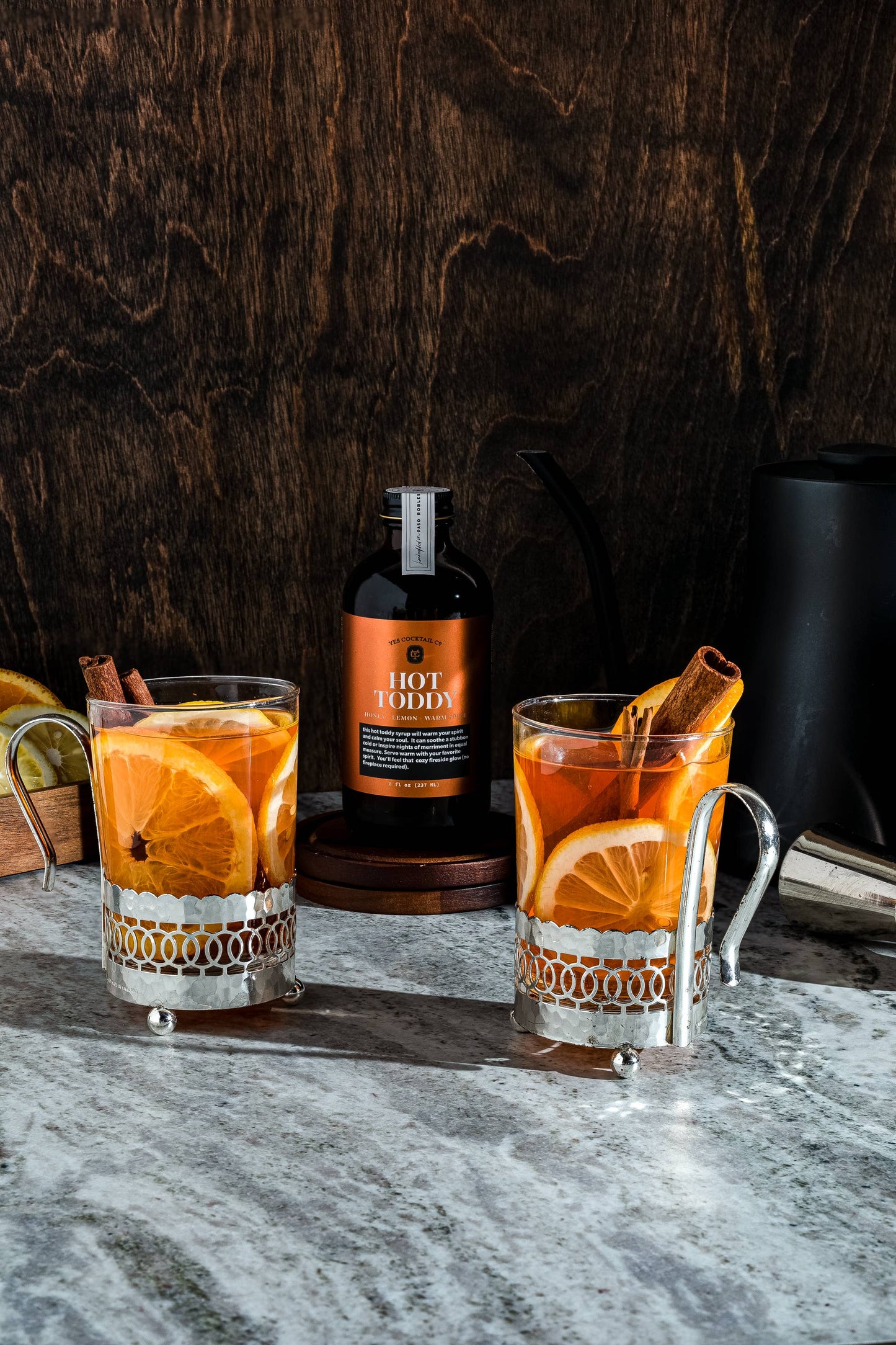 Two glasses of hot toddy with orange slices and cinnamon sticks on a marble surface, with a bottle of hot toddy in the background.