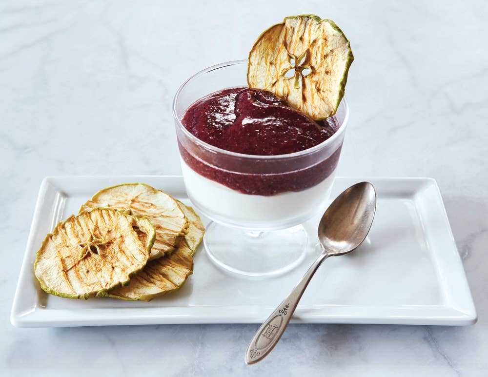 Glass of dessert with berry sauce, apple chips, and a spoon on a white plate.