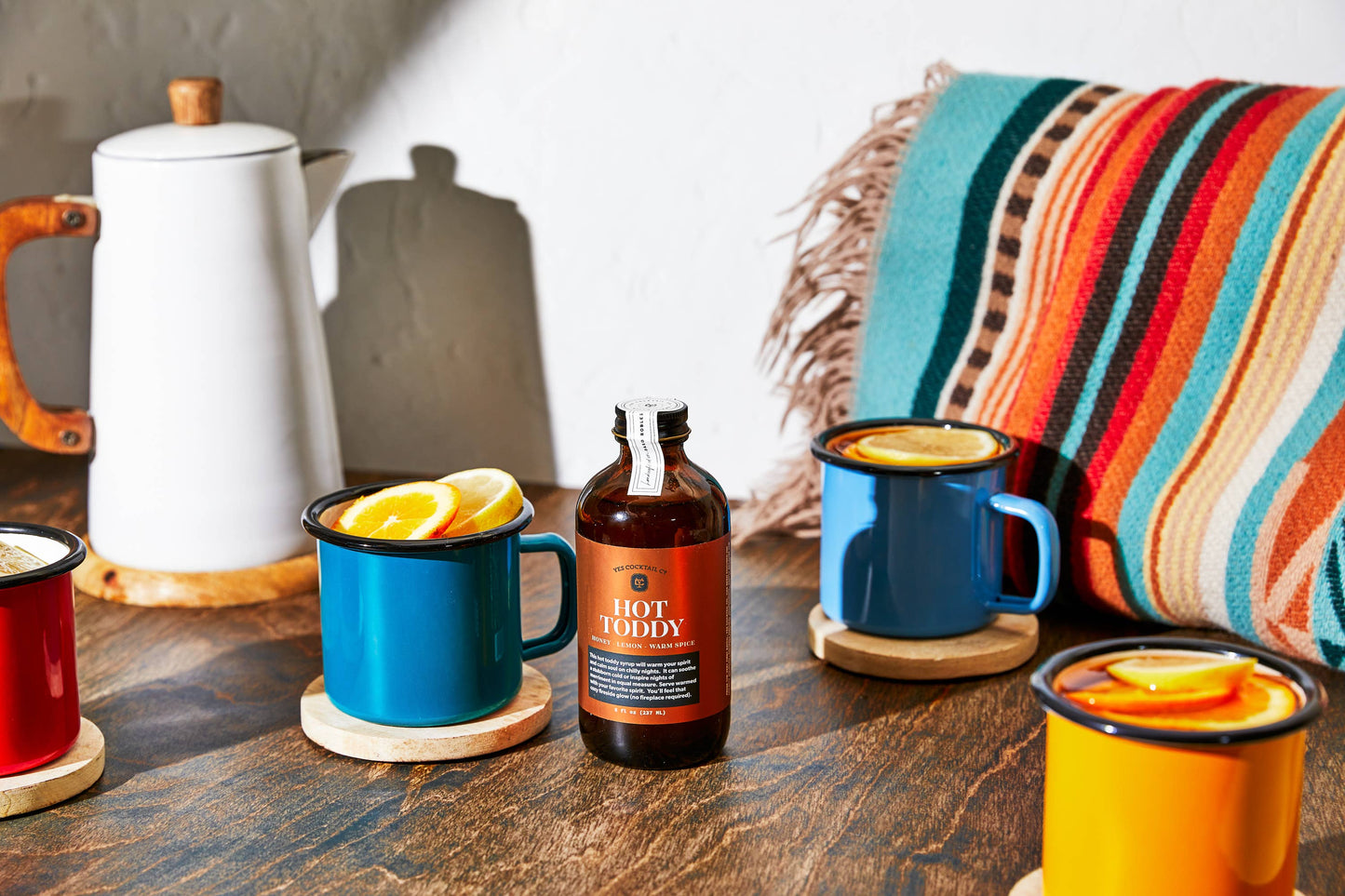 Hot toddy bottle with mugs of hot liquid on a wooden table with a colorful blanket in the background.