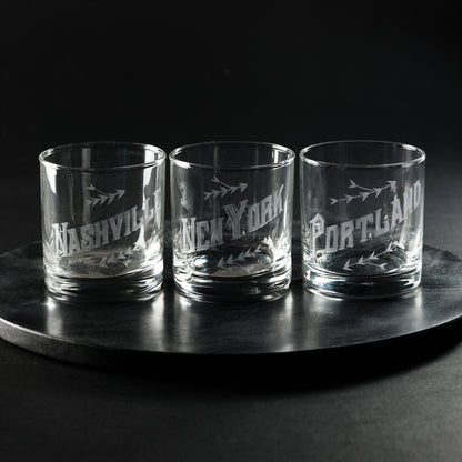 A set of three hand engraved low-ball glasses with city names inscribed on them, displayed on a circular dark surface.