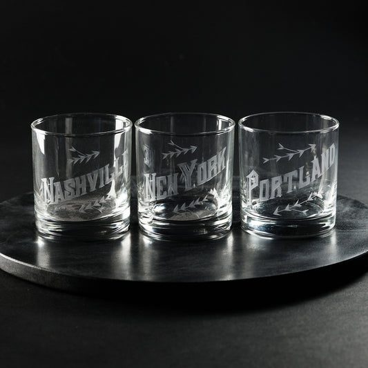 A set of three hand engraved low-ball glasses with city names inscribed on them, displayed on a circular dark surface.