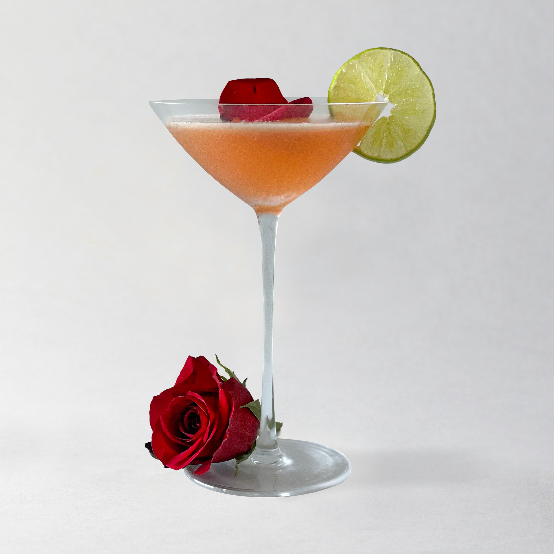 Cocktail in a martini glass with a red rose and lime wedge on a white background