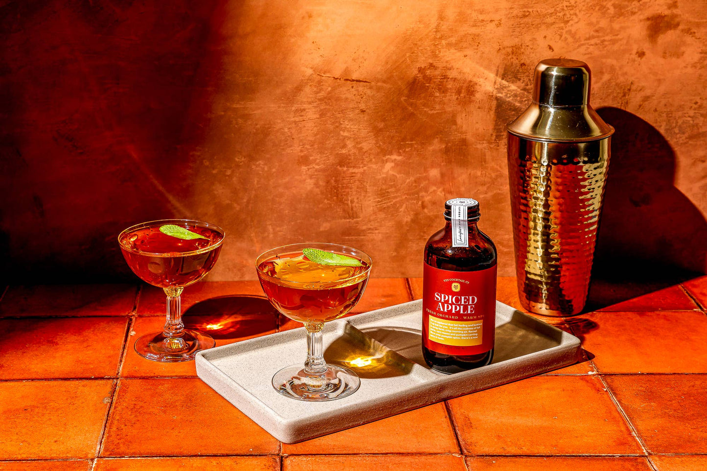Two cocktails in glasses with a bottle of spiced apple syrup and a gold shaker on a tray against an orange tiled wall.