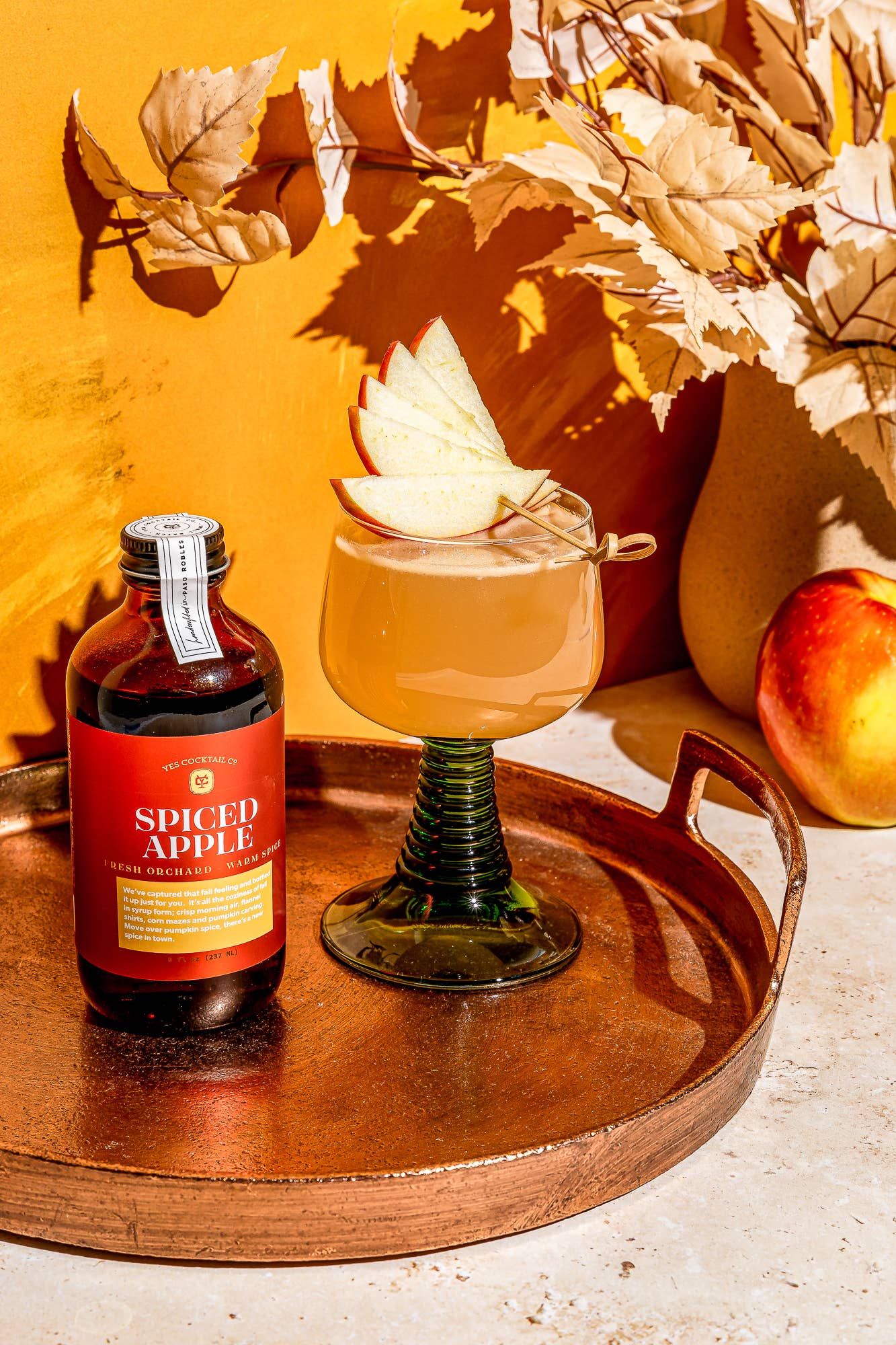 Spiced apple syrup bottle with a cocktail garnished with an apple slice on a wooden tray with autumn leaves and pumpkins.