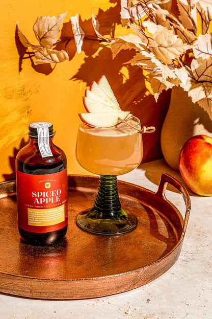 Spiced apple syrup bottle with a cocktail garnished with an apple slice on a wooden tray with autumn leaves and pumpkins.
